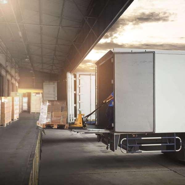 A driver unloads a pallet of boxes from a white semi-truck to a loading dock.