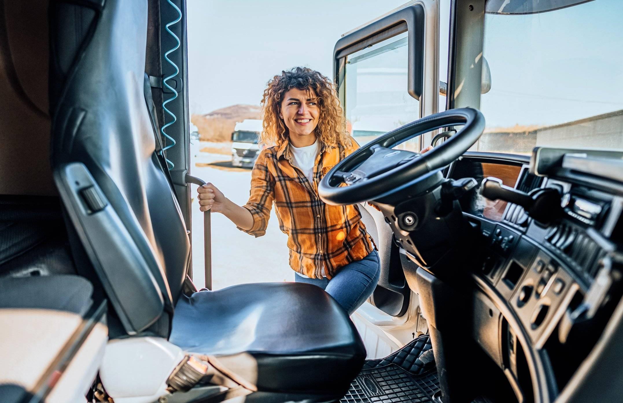 A female truck driver climbs into the cab of a semi truck while holding onto hand rails.