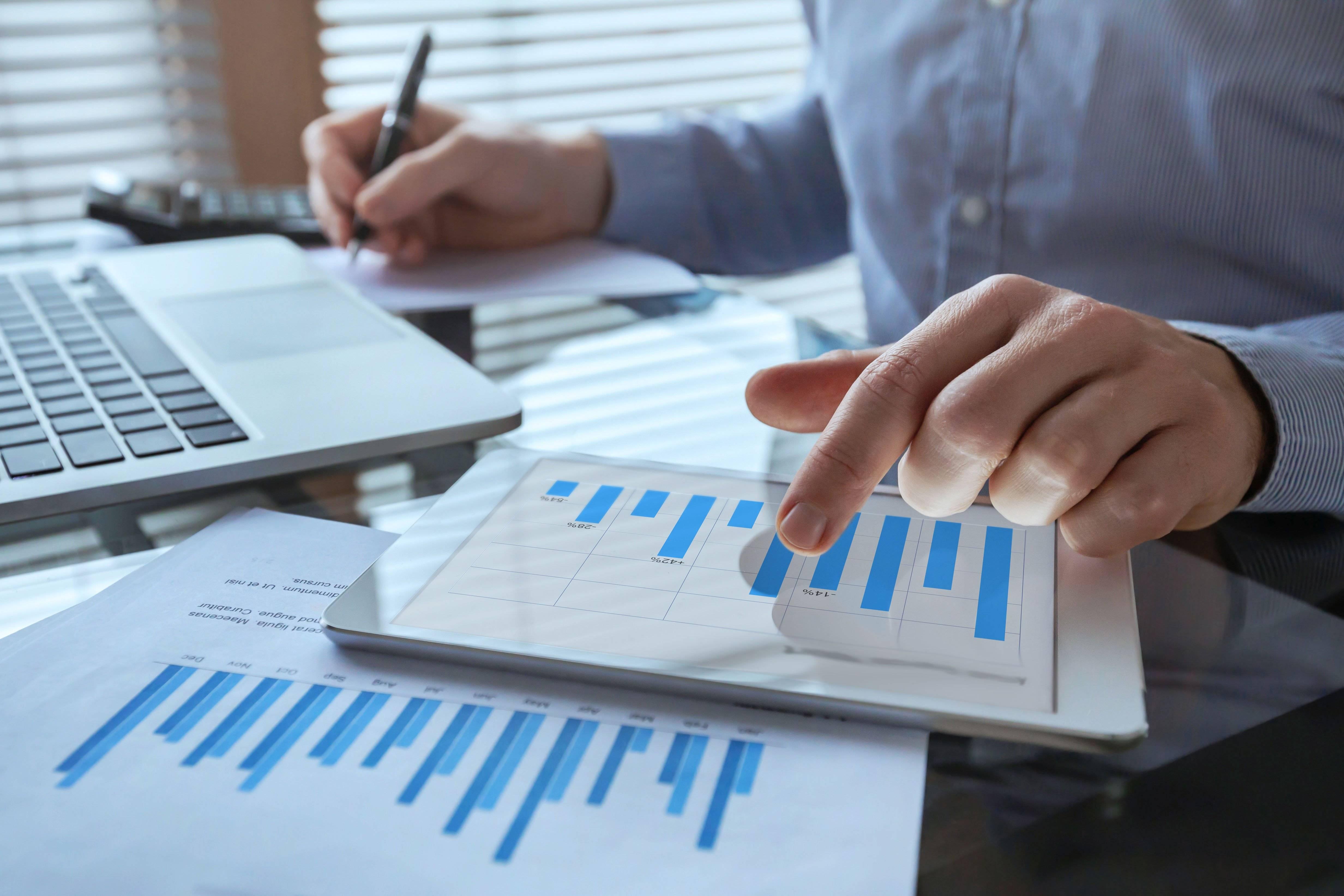 A hand points to a blue and white bar chart while the other hand holds a pen and takes notes by a laptop keyboard.