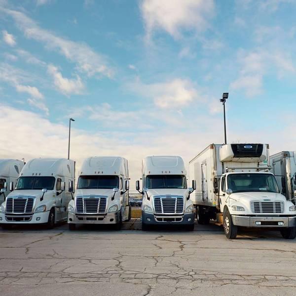 A line of white semi-trucks and box trucks parked in a lot against a blue cloudy sky.