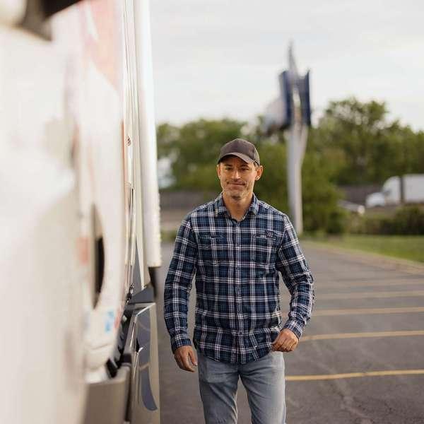 A man in a blue plaid flannel shirt walks in a parking lot next to a white semi-truck.