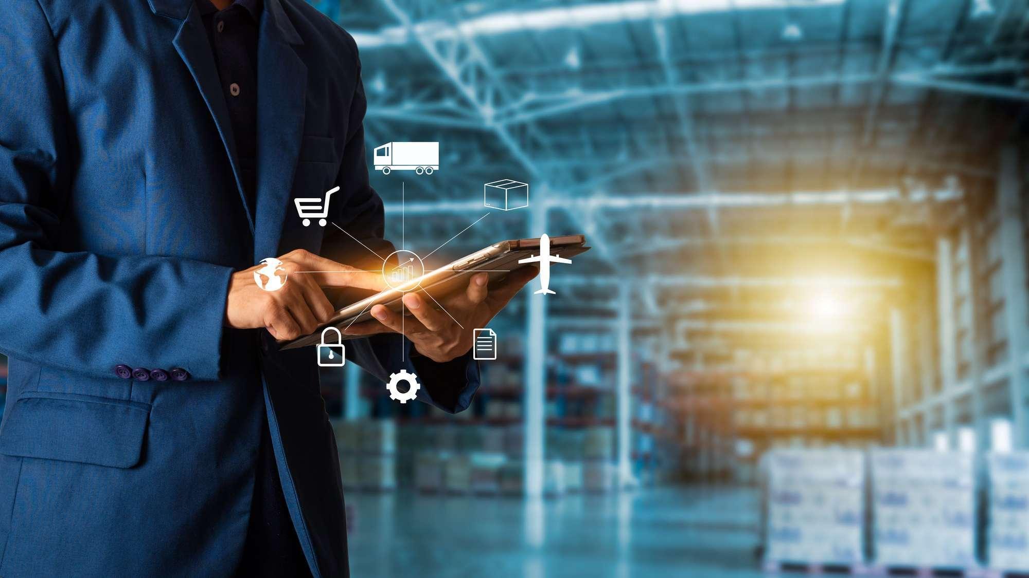 A man in a dark blue suit in a warehouse looks at a tablet device with a supply chain graphic overlayed on top.