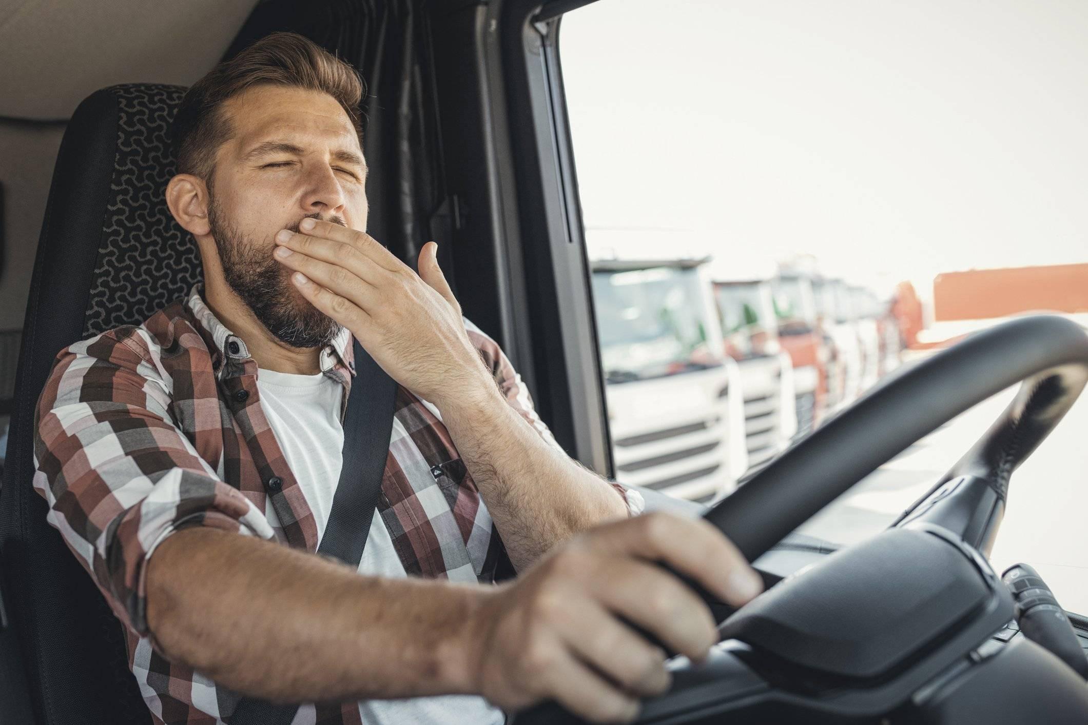 A man with a plaid shirt sits in a truck cab and covers his mouth with his hand while he yawns.
