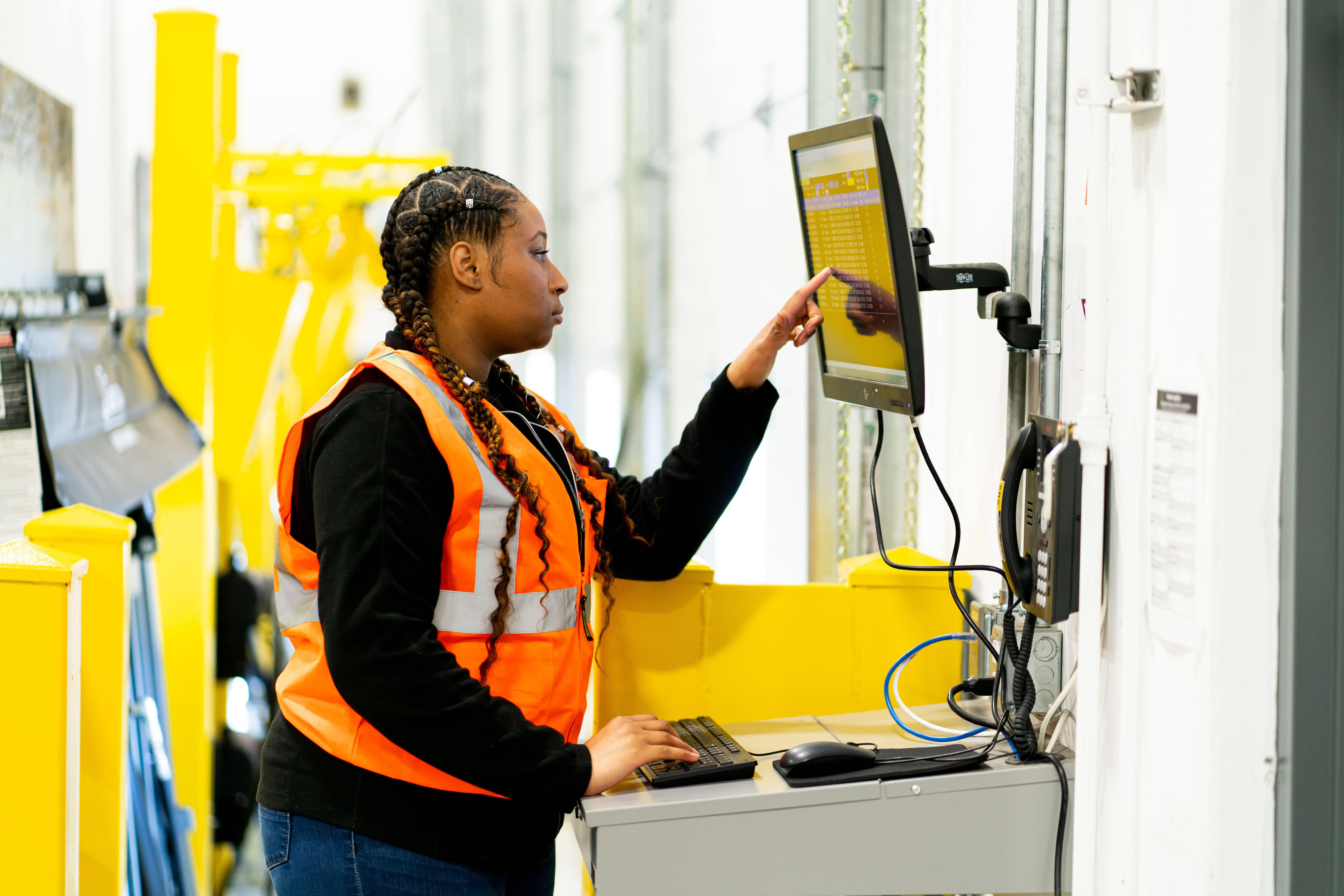 A woman wearing a safety vest works on a computer while in a warehouse.