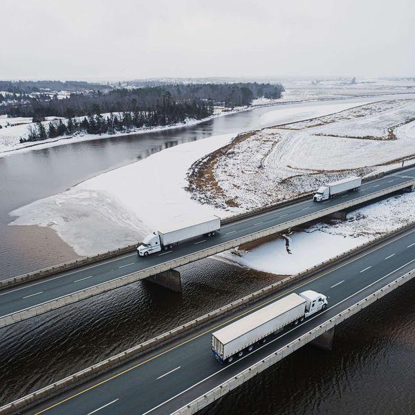 Three semi-trucks drive on a bridge across snowy land and water.