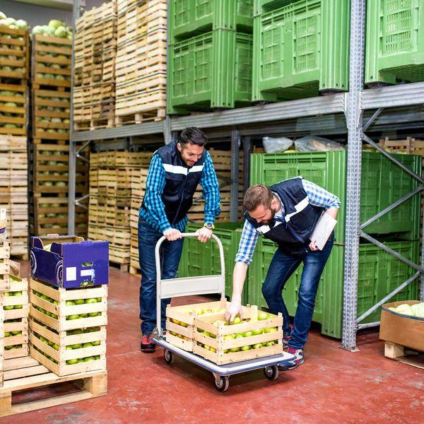 Two men pick through a crate of apples in a warehouse.