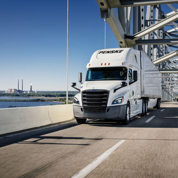 White Penske semi truck drives over a bridge next to water.